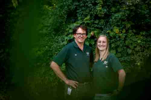 Two people in green John Ball Zoo shirts smile for the camera against a background of plants.